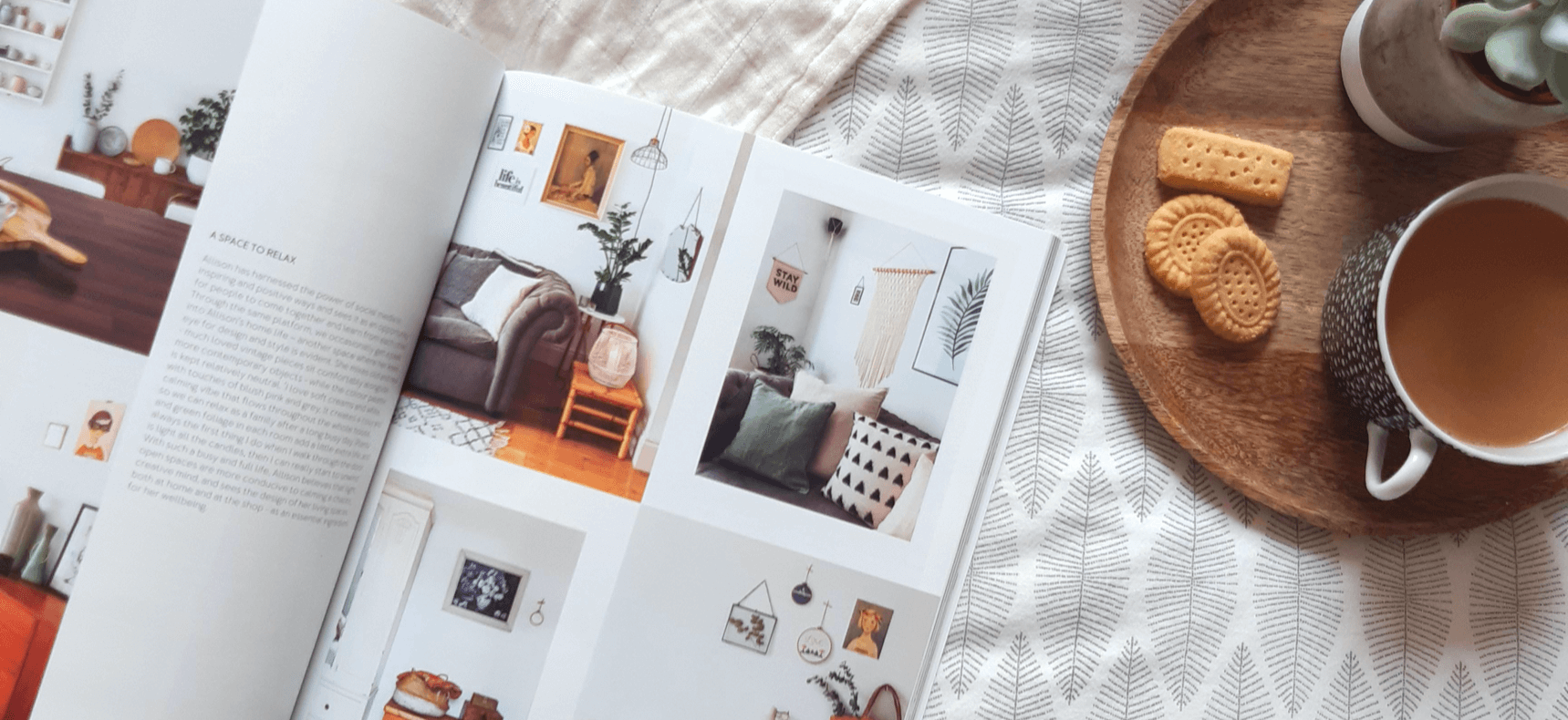 Open magazine showcasing interior design, cozy seating area, tea cup, and cookies on wooden tray.