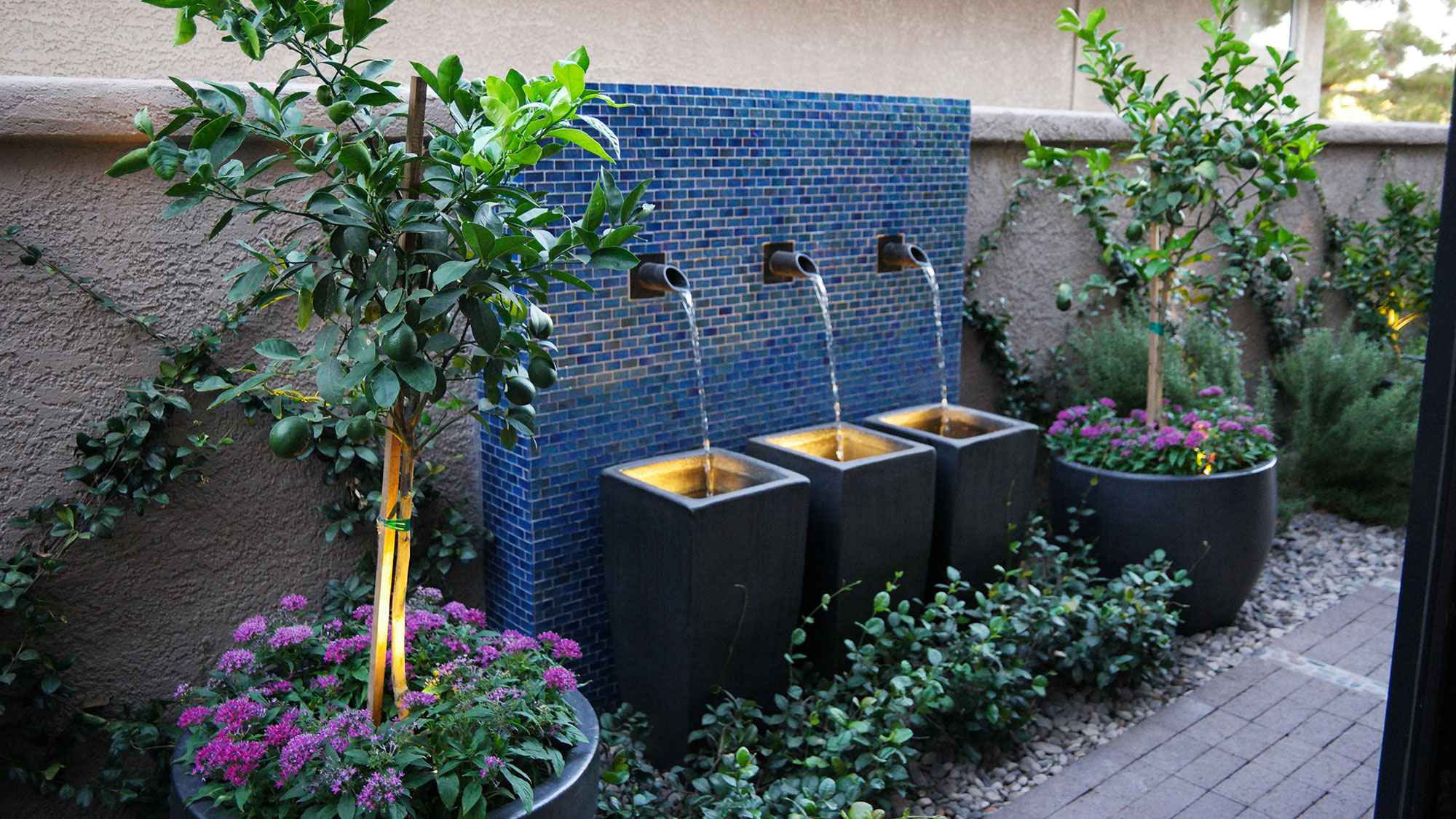 Three tiered water fountain with blue tile backdrop and lush potted plants.